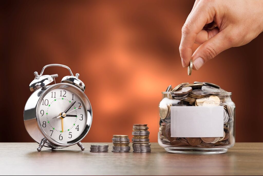 "Hand placing coin into a glass jar full of coins with a vintage alarm clock beside it, symbolizing time and money savings, investment, and financial growth."